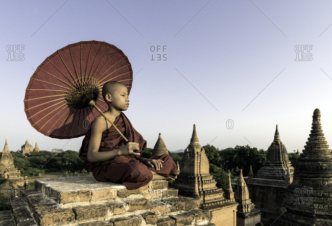 Young Buddhist monk sitting on a temple top holding a parasol