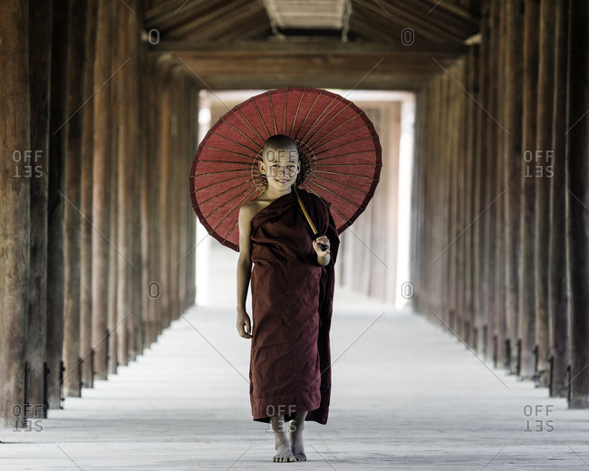 Young Buddhist monk walking along a temple complex corridor with parasol