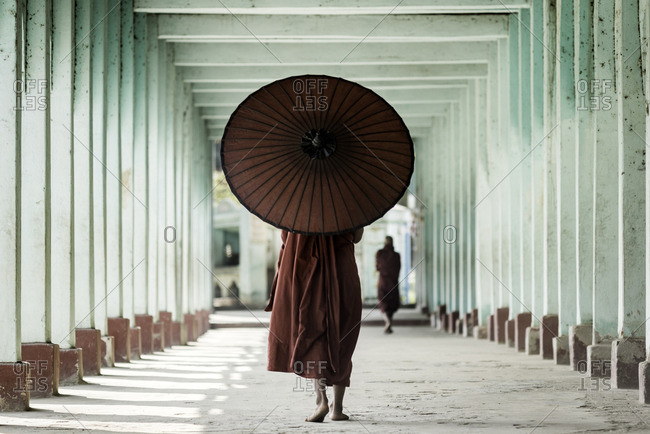 Young novice Buddhist monk walking along a temple complex corridor using a parasol