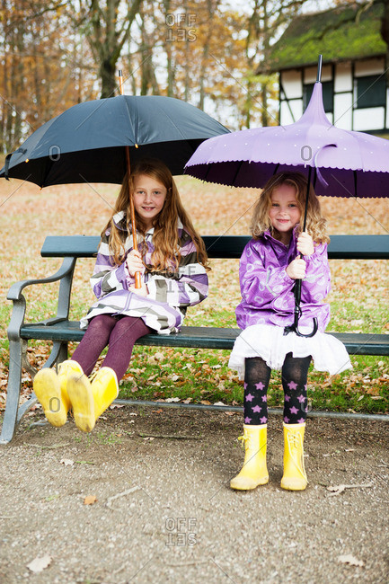 Girls in rain boots and umbrellas sitting in a bench in a park