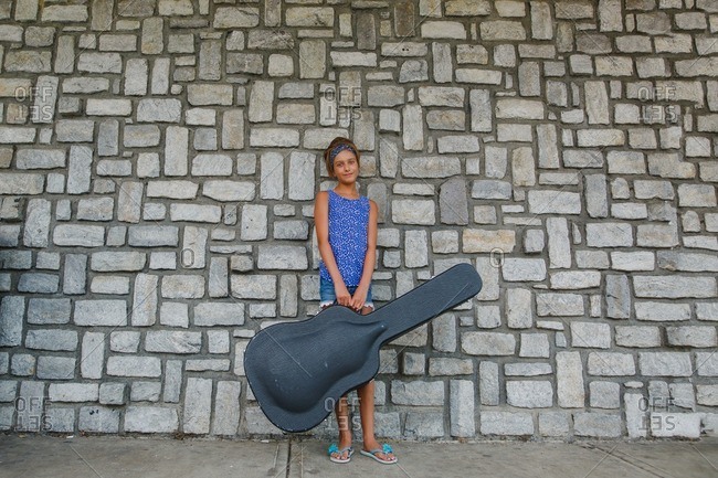 Tween girl standing against a stone wall holding a guitar case