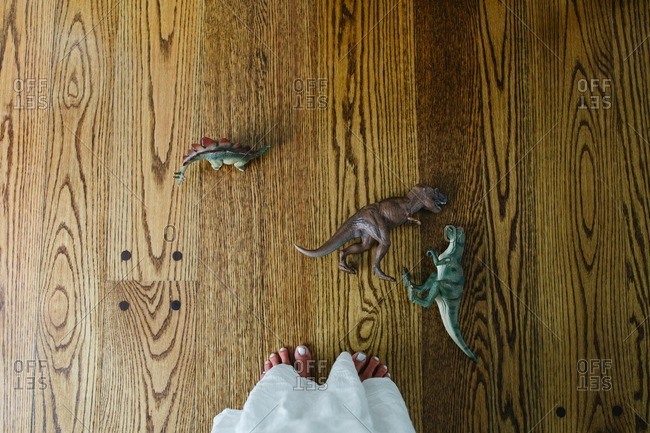 A woman with pedicured feet standing next to a group of toy dinosaurs on a wood floor