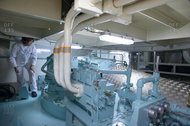 Engineer wearing ear defenders crouching under piped in ship\'s engine room
