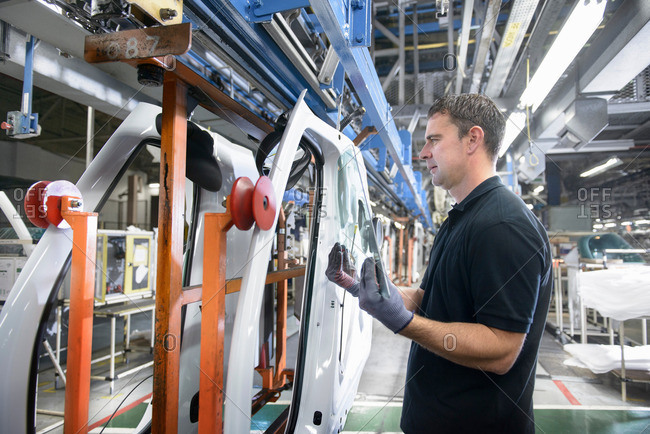 Car worker wearing protective gloves fitting sheet of glass into vehicle door on production line in car factory