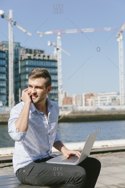 Ireland, Dublin, smiling young businessman sitting on bench with laptop telephoning with cell phone