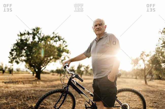Senior man with bicycle standing on a field at evening twilight