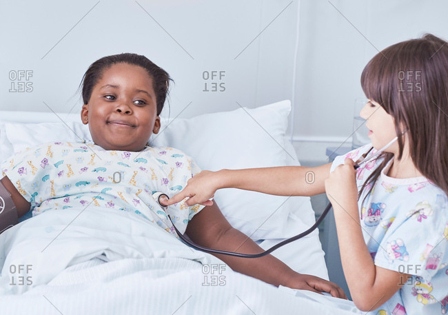 Girl patient using stethoscope on friend in bed on hospital children\'s ward