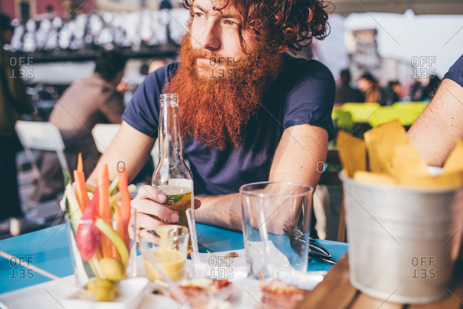 Young male hipster with red hair and beard drinking bottled beer at sidewalk bar