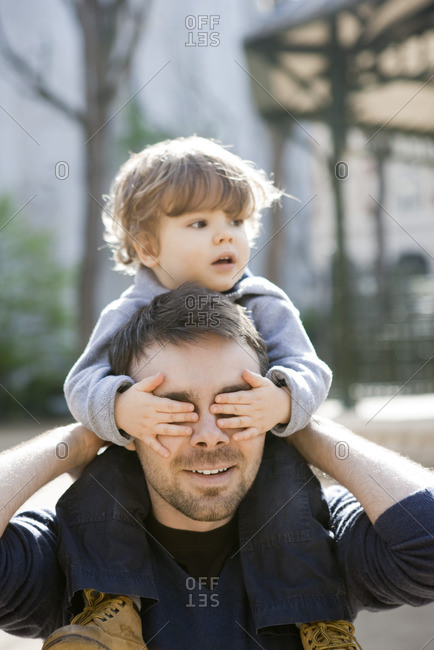 Father carrying toddler son on his shoulders, son covering father\'s eyes with his hands