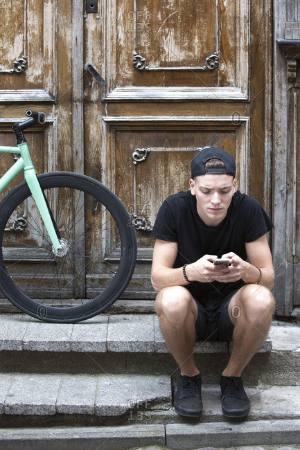 Young man using cell phone on stairs with parked bike at doors