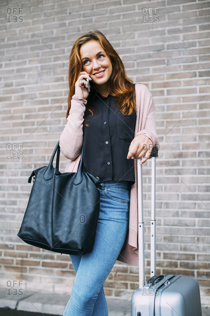 Smiling young woman with wheeled luggage and leather bag on the phone
