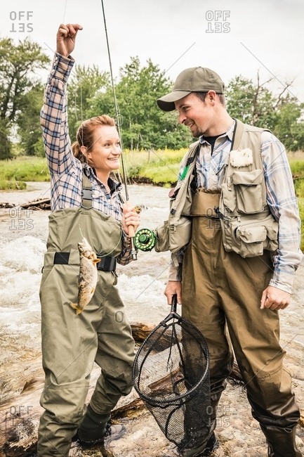 Man and woman standing in river wearing waders holding fishing rod smiling