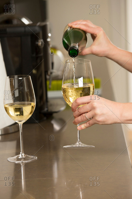 Hands of woman pouring glasses of white wine at kitchen counter