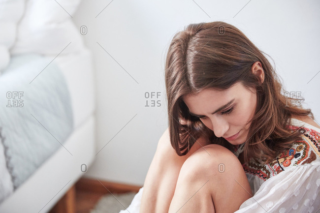 Young woman sitting on floor near bed hugging knees