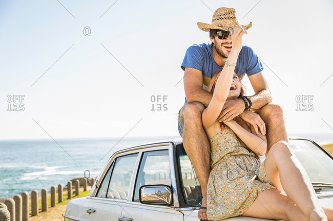 Mid adult couple sitting on top of car on coast road, Cape Town, South Africa