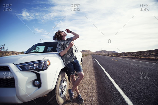 Young man leaning against car parked along the side of the road in the California desert