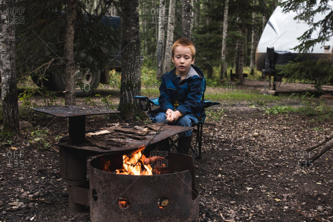 Boy sitting beside a campfire roasting a hot dog