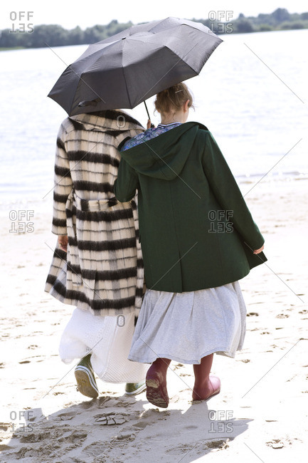 Back view of two friends walking side by side on the beach with an umbrella