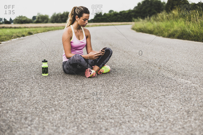 Sportive young woman sitting on street with cell phone and drinking bottle