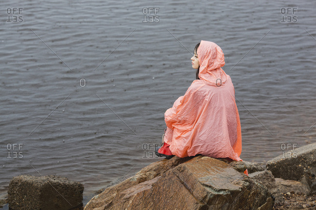 Thoughtful woman wearing raincoat sitting on rock at lakeshore during rainy season