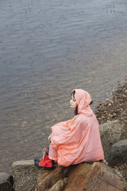 Thoughtful woman wearing raincoat sitting on rock at lakeshore in rainy season