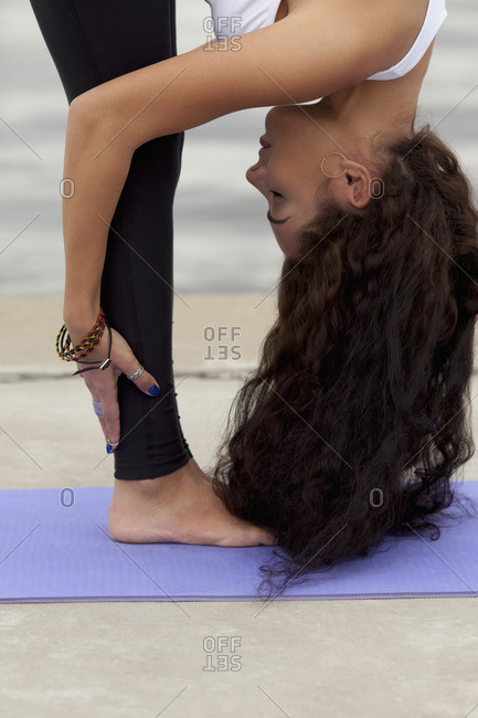 Side view of young woman practicing standing forward bend pose on exercise mat