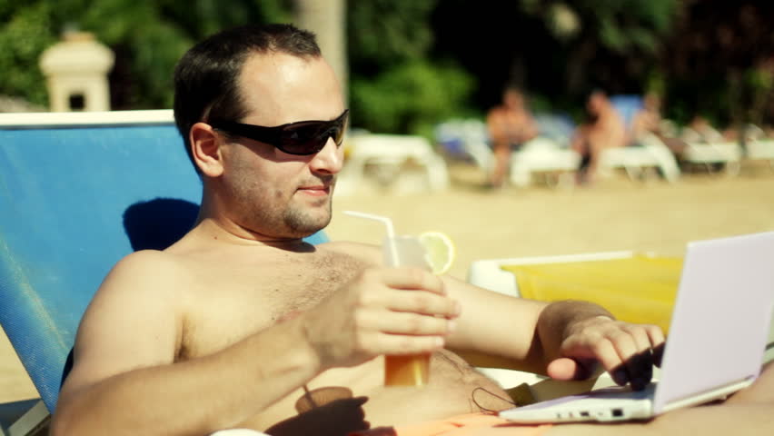 Handsome man lying on sunbed drinking cocktail and working on laptop 