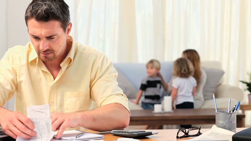 Casual man calculating his bills with his family in the background