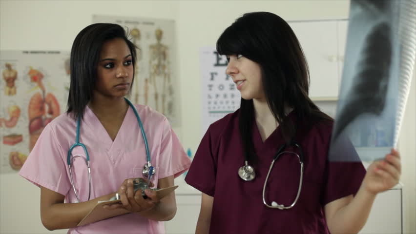 The camera dollies around keeping two young nurses or nursing interns in the frame as they discuss an x-ray film one of them is holding. Shallow depth of field. 