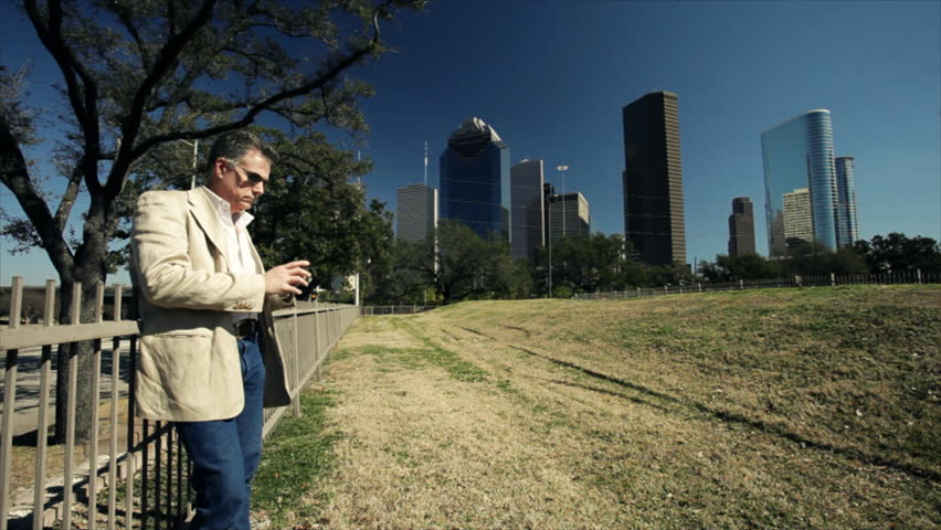 An attractive mature man standing against the fence of a downtown green space talks on his smart phone with metropolitan Houston looming in the background.