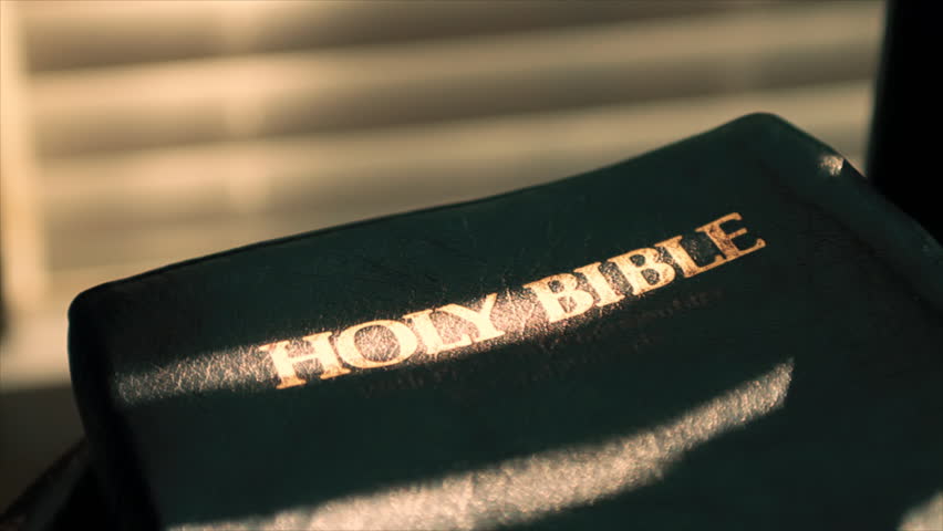 A woman's hand reaches down and picks up from a chair a Bible that is bathed in the sunbeams filtering through window shades.
