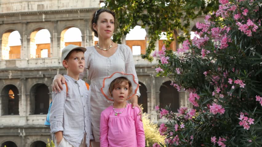 brunette woman says something to boy in white clothes and cap and girl in pink dress and  panama  in front of Colosseum in Rome