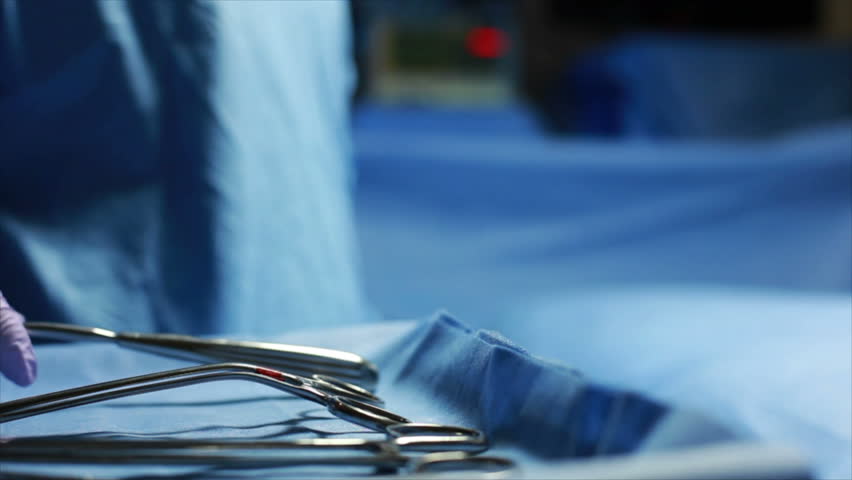 A surgical tool is handed to a surgeon in an operating room setting with out of focus blinking light in the background.