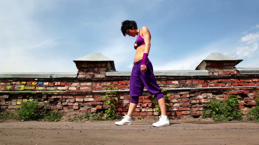 Woman in purple pants and a bra, performs modern dance, at background of small wall and sky with clouds