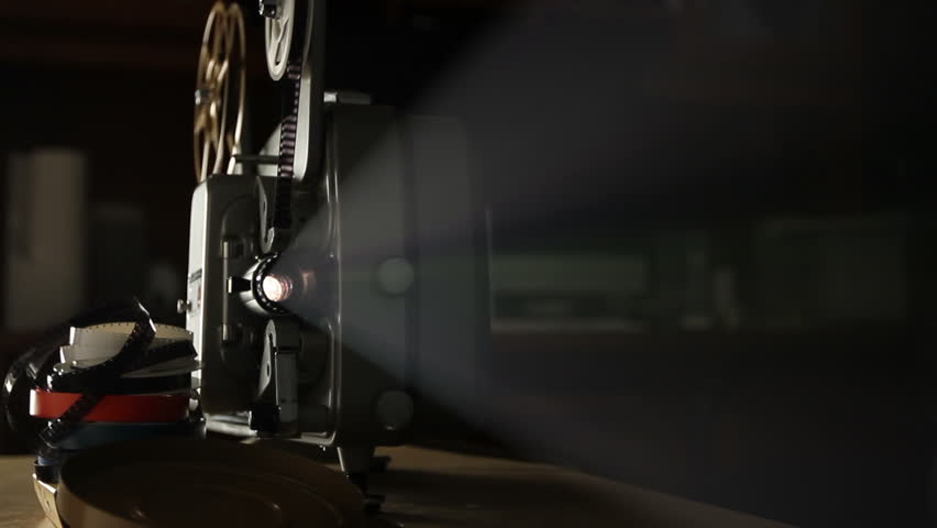 Front view of an old-fashioned antique Super 8mm film projector, projecting a beam of light in a dark room next to a stack of unraveled film reels. Includes projector audio.
