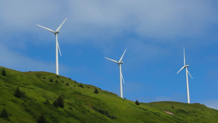 Three turbines spin fast, in unison, atop a lush, green hillside highlighted by cloud puffs in blue sky. 1080p
