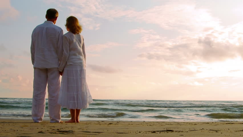 Grandparents and grandchildren enjoy a day at the beach.  Their activities include using a computer, walking on the beach, watching the sunset, having a drink, holding hands, and sharing an embrace.
