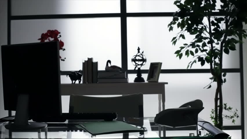 A businessman enters his very dimly lit office then sits down at his desk and starts going through a file. This creates a silhouette of the action due to backlighting.
