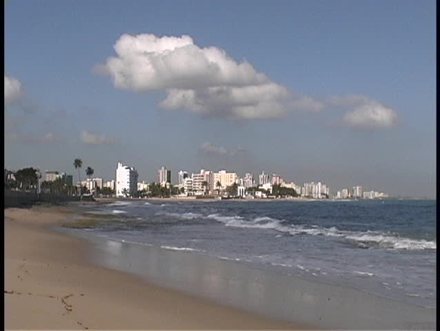 Waves break along a sandy beach in front of the San Juan skyline.