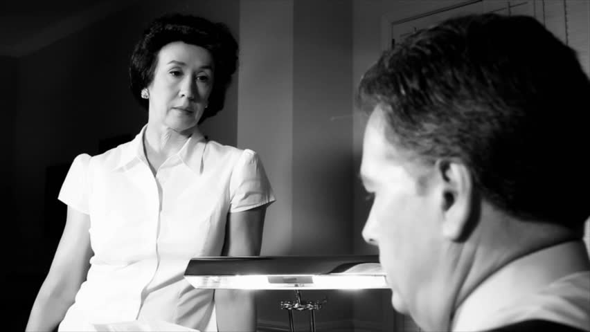 An annoyed then sympathetic or troubled woman dressed in 1940's style stands talking to a man sitting at a desk in front of her. Black and White. Film Noir Genre