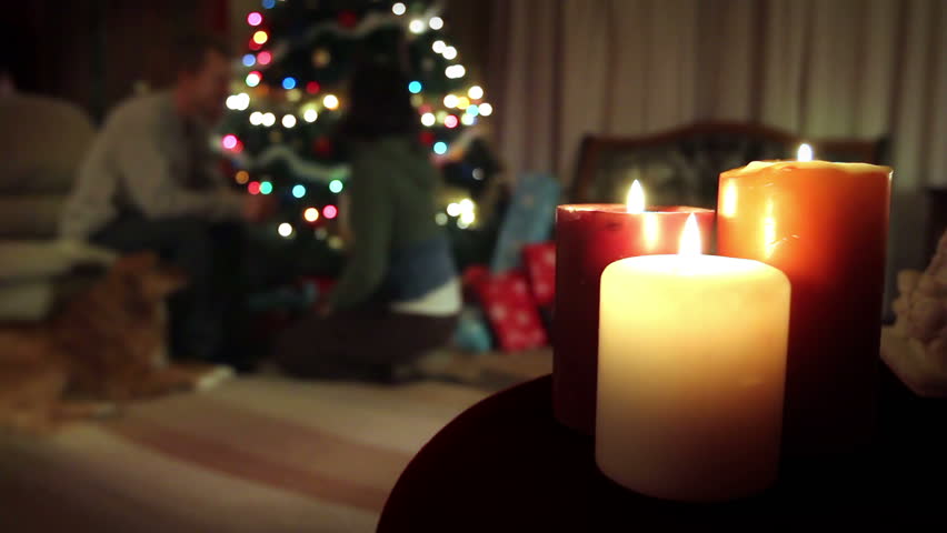 Christmas candles with a man, woman, and dog by the Christmas tree in the background. The woman unwraps a gift from the man, then hugs him.