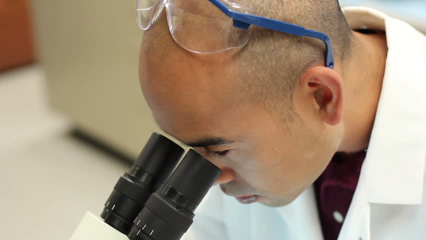 Asian researcher in a labcoat removes goggles, looks into a microscope and takes notes.