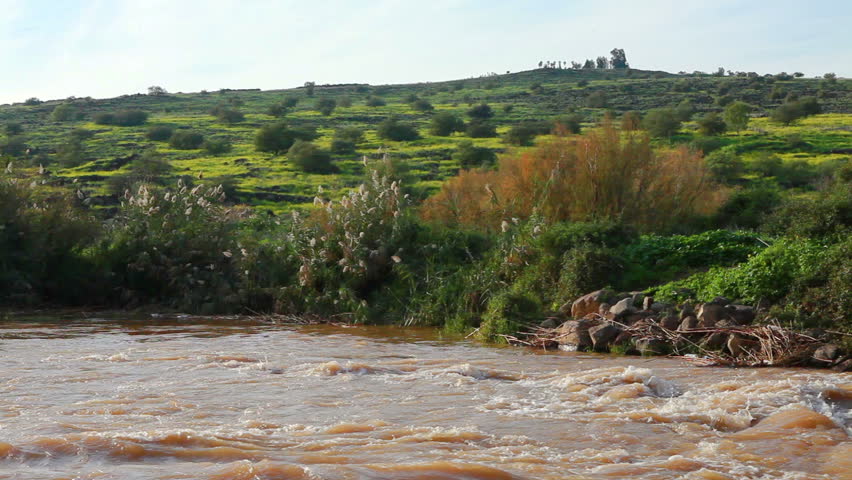 White water rapids on the reddish brown River Jordan in the Galilee of Israel.  In the background is a hillside covered with orange, yellow and green foliage.  