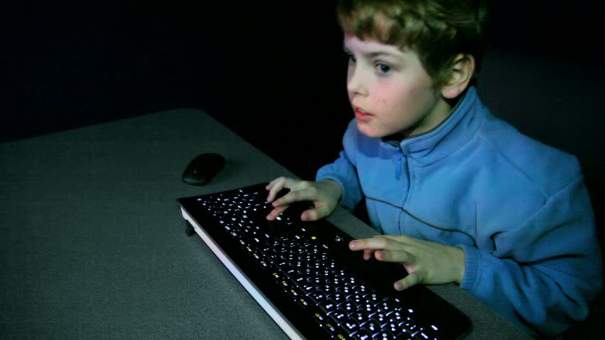 Young boy chews gum and plays video game with keyboard, mouse lay near on table in dark room