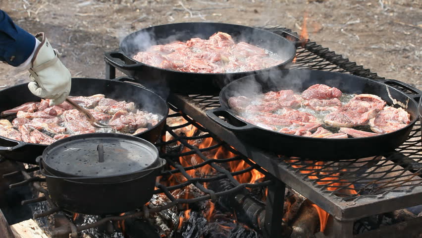 BBQ Lamb and mutton steaks and chops cooking in cast iron pans over an open outdoor fire. Cowboy and sheep camp bbq and party. Fresh meat with smoke and oil boiling near Dutch Oven. Close.