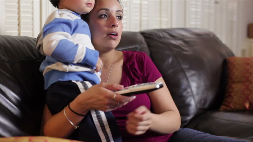 A lovely Hispanic mother with TV remote control in hand talks to her toddler about what they see on the television.