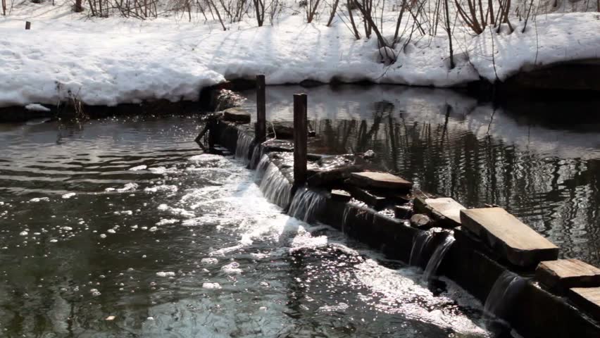 Snow lay on coast of river with small waterfall on dam