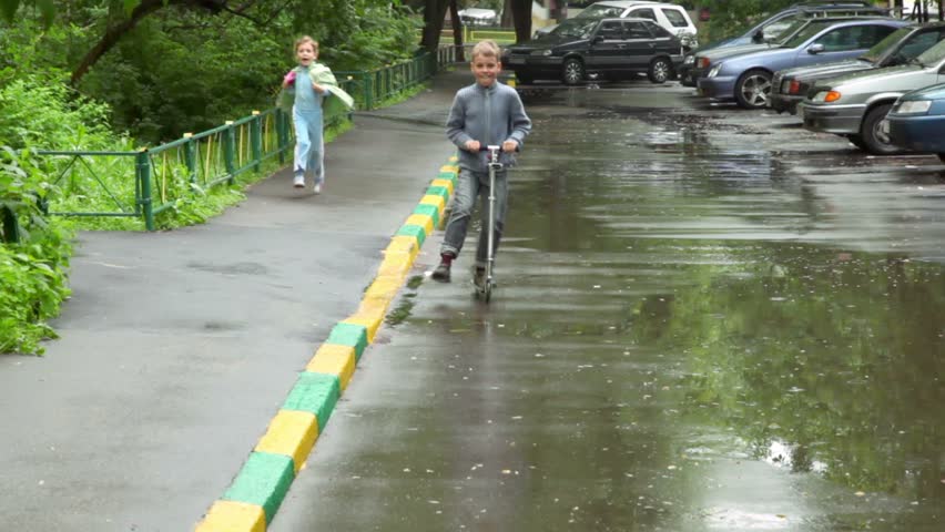 Little boy ride on scooter by asphalt and his sister run behind in rainy summer day