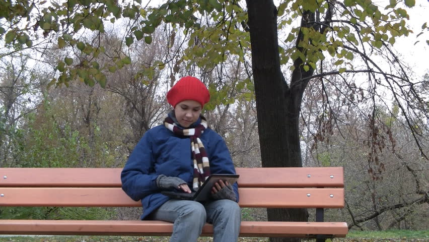 Pretty teenage girl sits on bench and works on netbook computer in autumn park