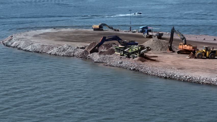 Truck rides and cranes work at construction site at shore near water where boat floats